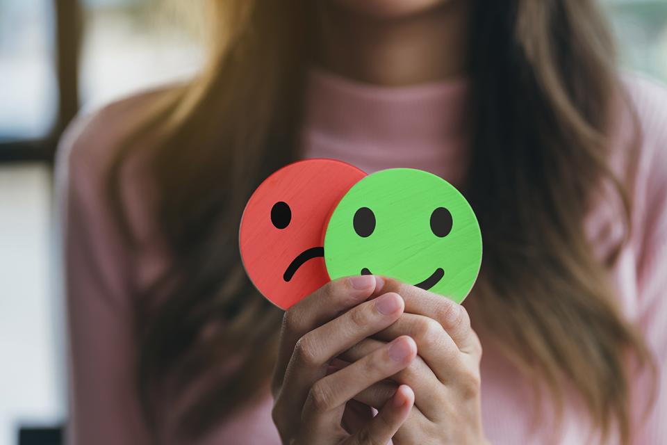 Young woman holding up smiley face in front of a frown face, depicting mental health for Mental Health Awareness month. 